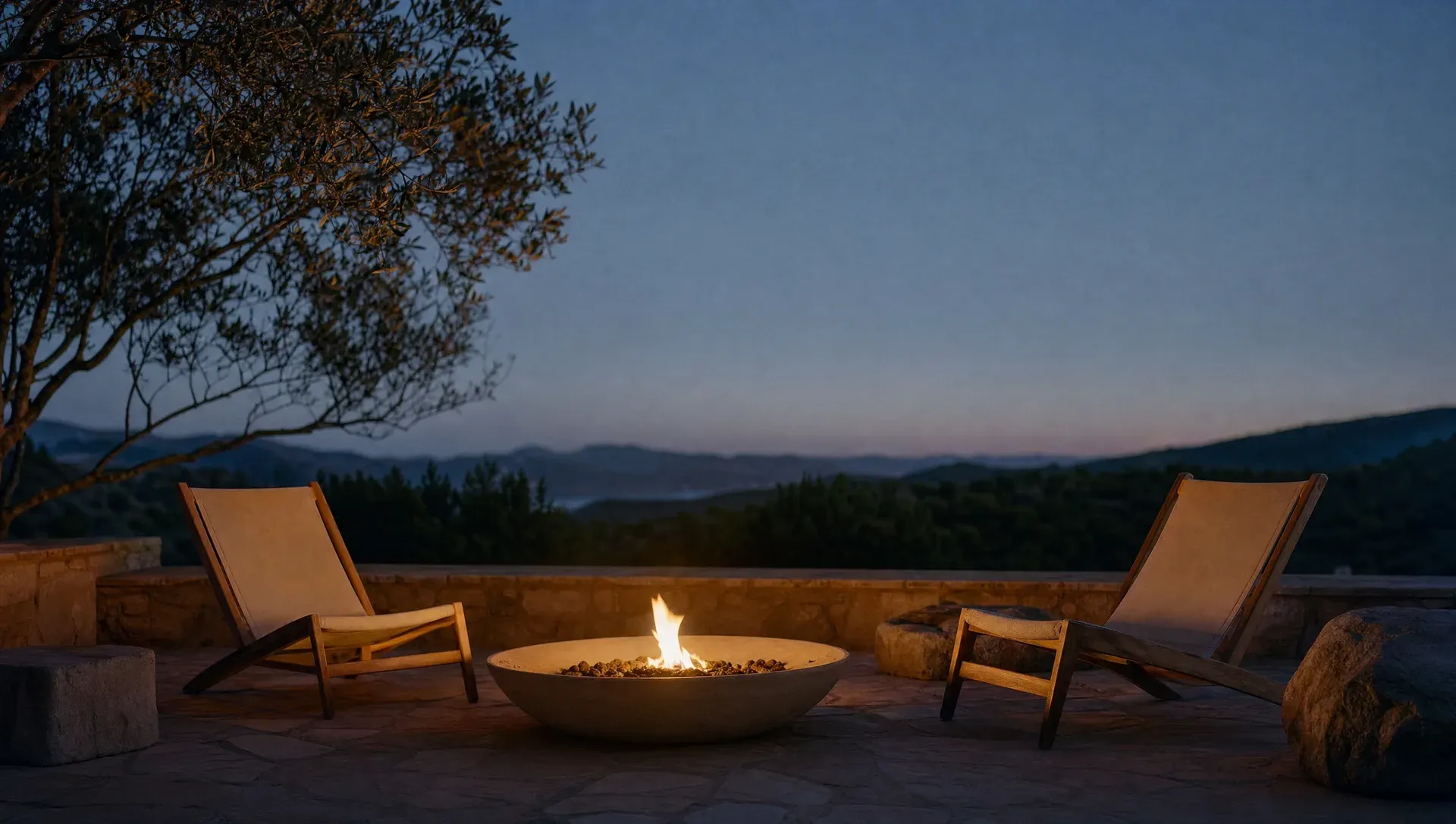 A fire pit glowing on a stone terrace at dusk, an olive tree and hills beyond.
